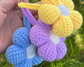 May include: Three crocheted flower keychains in yellow, purple, and blue. Each flower has a white center.