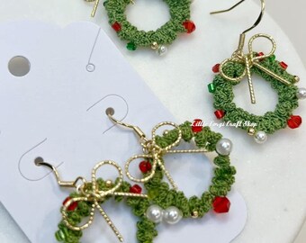 May include: Close-up of three pairs of Christmas wreath earrings. Each earring features a green crocheted wreath adorned with red, green, and white beads, and a gold-colored bow. The earrings are displayed on a white card.