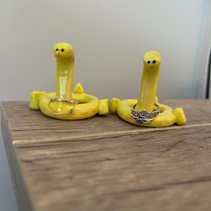 Pu&ograve; includere: Due stravaganti portaanelli in ceramica gialla a forma di vermi sorridenti. Ogni supporto ha una base circolare e un corpo di verme verticale. Uno tiene un delicato anello d'oro con un piccolo ciondolo, l'altro un anello d'argento con un motivo a corona. Posizionati su una superficie di legno.