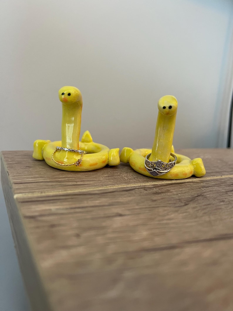 Pu&ograve; includere: Due stravaganti portaanelli in ceramica gialla a forma di vermi sorridenti. Ogni supporto ha una base circolare e un corpo di verme verticale. Uno tiene un delicato anello d'oro con un piccolo ciondolo, l'altro un anello d'argento con un motivo a corona. Posizionati su una superficie di legno.
