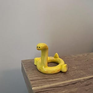Pu&ograve; includere: Statuetta a forma di serpente in ceramica gialla con corpo curvo e testa eretta. Il serpente ha piccoli occhi neri e segni arancioni. La statuetta &egrave; esposta su una superficie di legno.