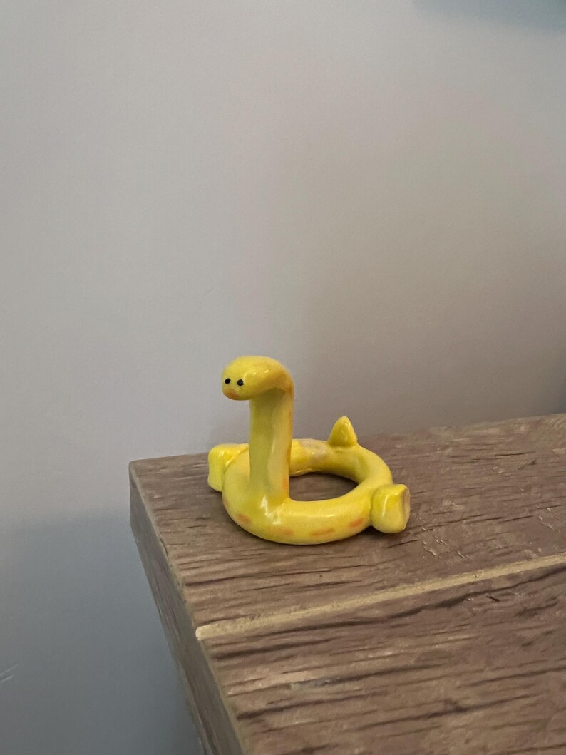 Pu&ograve; includere: Statuetta a forma di serpente in ceramica gialla con corpo curvo e testa eretta. Il serpente ha piccoli occhi neri e segni arancioni. La statuetta &egrave; esposta su una superficie di legno.