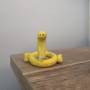 Puede incluir: Un caprichoso portaanillos de cerámica amarilla con forma de gusano, con un anillo. El soporte tiene una base circular, un cuerpo elevado en forma de gusano y una pequeña cabeza. El anillo es plateado con un diseño floral.