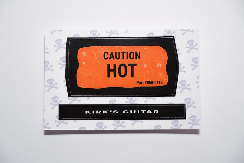Może przedstawiać: Czarno-pomarańczowa naklejka z tekstem "CAUTION HOT" i "Part #850-0113". Pod naklejką znajduje się czarna naklejka z tekstem "KIRK'S GUITAR" na białym tle z wzorem małych szarych czaszek i skrzyżowanych kości.