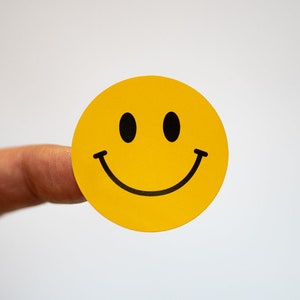 Könnte beinhalten: Ein gelber Smiley-Aufkleber mit schwarzem Rand. Der Aufkleber wird von einem Finger gehalten.