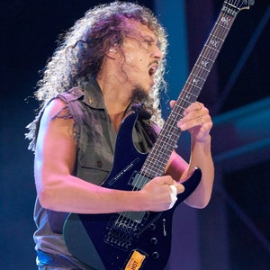 Może przedstawiać: Mężczyzna gra na czarnej gitarze elektrycznej z białym pickguadem. Gitara ma na korpusie czarno-biały motyw czaszki i napis "Kirk's Guitar". Gitara ma r&oacute;wnież naklejkę z napisem "Dunlop HOT".