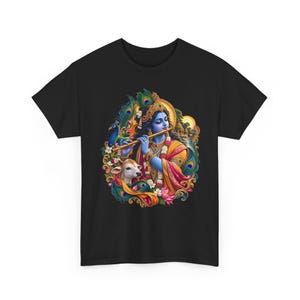 Könnte beinhalten: Ein schwarzes T-Shirt mit einer farbenfrohen, detaillierten Grafik einer blauhäutigen Figur, die eine goldene Flöte spielt. Die Abbildung zeigt ein weißes Kalb, Pfauenfedern, Lotusblüten und eine leuchtende Sonne, umrahmt von kunstvollen Mustern.