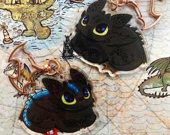 Puede incluir: Dos llaveros acrílicos con un dragón negro de ojos grandes y un diseño lindo. Los llaveros tienen cierres de color oro rosa. El fondo es un mapa con ilustraciones de dragones y otros elementos de fantasía.