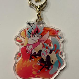 May include: A colorful acrylic keychain featuring a cartoon character with blue and pink hair, wearing a pink shirt with a white heart, and a blue and white patterned skirt. The character is sitting on a red, orange, and blue splatter background.