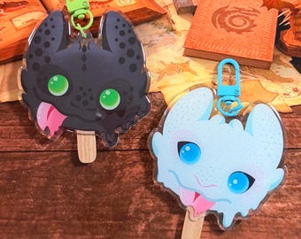 Puede incluir: Dos llaveros de acrílico con forma de helado de palo que presentan cabezas de dragón estilizadas. Uno es negro con ojos verdes y un clip verde lima, el otro es azul claro con ojos azules y un clip azul claro. Ambos tienen una lengua rosa y un palo.