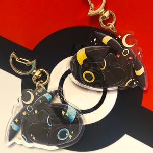 Puede incluir: Dos llaveros de personajes Pokémon negros y azules con cierres de metal dorado. Los personajes están acurrucados con los ojos cerrados. Los llaveros están sobre un fondo rojo, blanco y negro.