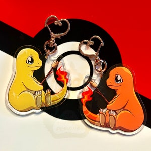 May include: Two clear acrylic keychains featuring cartoon versions of the Pokemon Charmander and Squirtle. Both Pokemon are holding a stick with a marshmallow on the end, as if they are roasting it over a fire. The keychains are on a red and black background.
