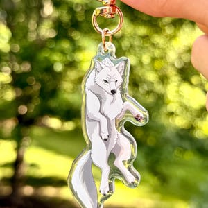 Puede incluir: Un llavero de acrílico transparente con una ilustración estilizada de un lobo blanco con sombreado gris. El lobo aparece en una pose reclinada con la cola curvada hacia abajo. El dije está sujeto a un broche de langosta giratorio de metal dorado.