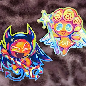 May include: Two colorful stickers featuring cartoon characters. One is a devil with horns, wings, and a candy cane. The other is an angel with wings and a star-topped wand. Both have vibrant colors and a playful design.
