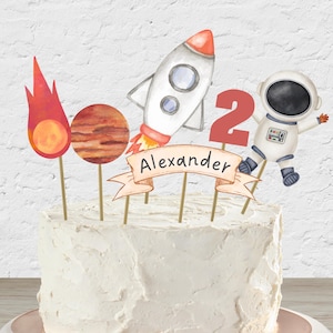 Op de afbeelding: Een witte taart versierd met een raket, een planeet, een zon, een astronaut en een banner met de tekst "Alexander" met het nummer "2" erop.