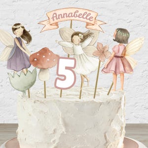 Op de afbeelding: Een witte taart met een roze en wit cijfer 5, een banner met de tekst "Annabelle" en drie feeënfiguren met vleugels. De feeën dragen paarse, witte en roze jurken.