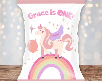 May include: White party bag with a pink border and the text "Grace is ONE!". The bag features a cartoon unicorn with pink hair and wings, standing on a rainbow, with a pink balloon. Stars and sparkles are scattered around the unicorn.