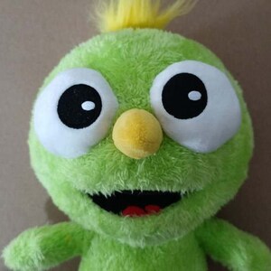 May include: A green plush toy with a yellow tuft of hair, large white eyes with black pupils, and a yellow beak. The toy has a wide, open mouth with a red tongue.