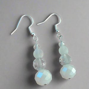 May include: A pair of silver dangle earrings with three white glass beads. The bottom bead is faceted, the middle bead has a snowflake design, and the top bead is frosted.