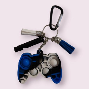 May include: A blue and black silicone fidget toy shaped like a video game controller. The toy has a silver key ring attached to it with a black whistle, a white tassel, and a blue tassel.