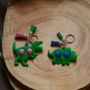 May include: Two green dinosaur-shaped keychains with colorful push-button fidget toys. One dinosaur is a Tyrannosaurus Rex with pink buttons and the other is a Triceratops with blue buttons. Both keychains have a silver ring and a green tassel.