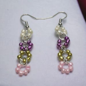 May include: A pair of handmade dangle earrings. Each earring features a silver hook, with a series of beaded rings in white, purple, gold, and pink. The earrings are made with small beads and silver connecting rings.
