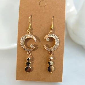 May include: Gold-toned dangle earrings featuring a crescent moon design encrusted with clear crystals and a small star. The earrings have a three-bead dangle, with a star-shaped bead, a faceted bead, and a round bead. The earrings are displayed on a brown card.