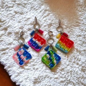May include: Four colorful pop it keychain toys with tassels. The keychains are square shaped and have a variety of colors including blue, green, yellow, red, and white.