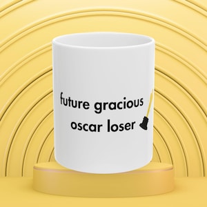 May include: White ceramic mug with the text "future gracious oscar loser" in black. A yellow and black illustration of an Oscar statue is on the right side of the mug. The mug is on a yellow pedestal.