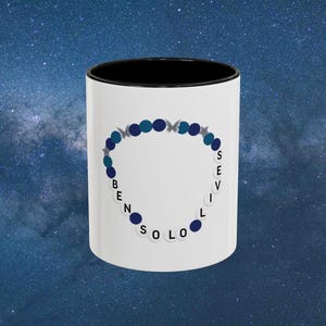 Peut inclure: Mug en céramique blanche avec intérieur noir. Il présente un motif de bracelet composé de perles bleues et d'étoiles argentées, avec les mots "BEN SOLO SEVIL" autour du motif.