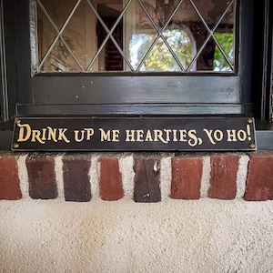 May include: A black wooden sign with gold lettering that reads "Drink Up Me Hearties, Yo Ho!" The sign is mounted above a brick ledge.
