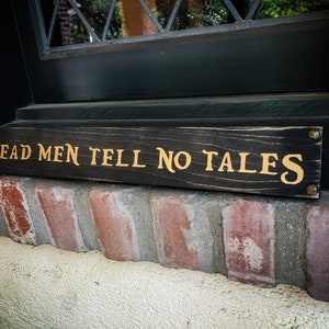 May include: A black wooden sign with gold lettering that reads "Dead Men Tell No Tales".