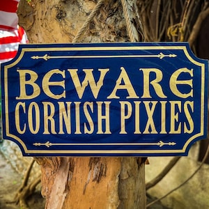 May include: A blue wooden sign with gold lettering that reads "Beware Cornish Pixies". The sign is hanging on a tree trunk.