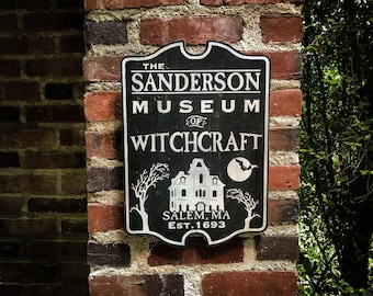 May include: A black and white sign for The Sanderson Museum of Witchcraft, mounted on a brick wall. The sign features a stylized illustration of a house, a full moon with a bat, and the text "Salem, MA EST. 1693."