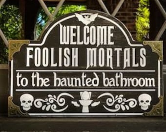 Könnte beinhalten: Schwarzes Holzschild mit goldenen Akzenten, mit dem Text "Welcome Foolish Mortals to the haunted bathroom". Das Schild hat ein Fledermaus-, Totenkopf- und Toiletten-Symbol.