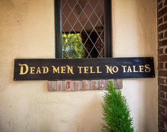May include: A rectangular wooden sign with the phrase "DEAD MEN TELL NO TALES" in gold lettering. The sign is mounted on a brick ledge below a window with a diamond-patterned pane. A small evergreen bush is in the foreground.