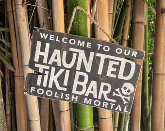 May include: A black wooden sign with white text that reads "Welcome to our Haunted Tiki Bar, Foolish Mortal". The sign has a skull and crossbones graphic and is hanging from a rope in front of a bamboo forest.
