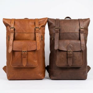 Può includere: Due zaini in pelle posizionati uno accanto all'altro. Lo zaino a sinistra è marrone chiaro, quello a destra è marrone scuro. Entrambi presentano un design roll-top, due cinghie verticali con fibbia, una tasca centrale con fibbia e cerniere laterali.