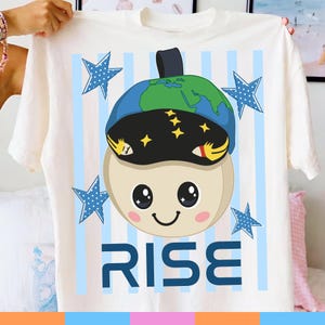 May include: Off-white t-shirt featuring a cartoon character wearing a globe-shaped hat and a black visor with stars. The shirt has blue stars and vertical blue stripes. The word "RISE" is printed in blue block letters.