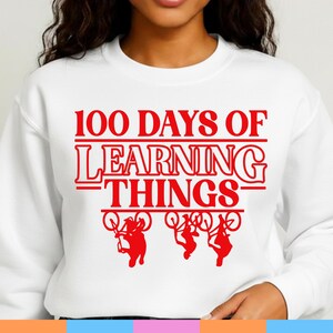 May include: White sweatshirt featuring the red text "100 DAYS OF LEARNING THINGS" and silhouettes of figures on bicycles. The design is centered on the front of the long-sleeved top.