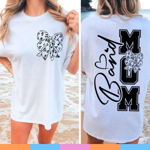Puede incluir: Camiseta blanca con un diseño de lazo negro lleno de notas musicales en la parte delantera. La parte trasera de la camiseta muestra las palabras "Band Mom" en negro, con un corazón y un lazo.