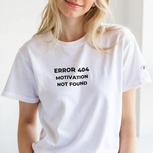 Op de afbeelding: Wit T-shirt met de tekst "ERROR 404 MOTIVATION NOT FOUND" in het zwart. Het shirt heeft korte mouwen en een klein ontwerp op de rechter mouw. Het model draagt een bruine broek.