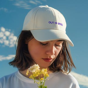 Pode incluir: Boné de beisebol branco com o texto "OUT OF OFFICE" em roxo. Uma pessoa segura um pequeno buquê de flores amarelas e brancas. O fundo é um céu azul brilhante com nuvens dispersas.