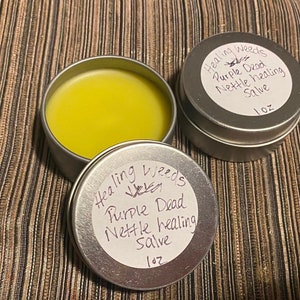 Puede incluir: Dos latas plateadas de bálsamo curativo de ortiga muerta púrpura, cada una etiquetada con el nombre del producto y "Healing Weeds". El bálsamo es de color amarillo brillante.