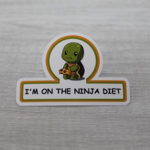 May include: A white sticker with a green turtle cartoon holding a slice of pizza. The sticker has a green and orange border and the text "I'm on the ninja diet".