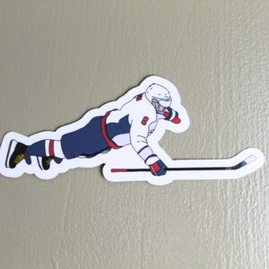 May include: A sticker featuring a hockey player in a white, navy blue, and red uniform, mid-slide. The player wears a helmet and gloves, with a hockey stick extending to the right. The number 8 is visible on the jersey.