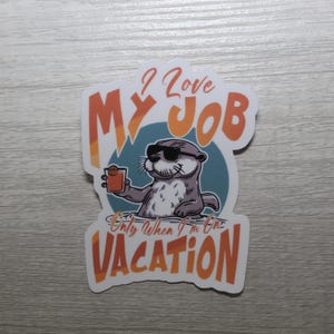 May include: A sticker with a cartoon otter wearing sunglasses and holding a drink. The text on the sticker reads "I love my job only when I'm on vacation."