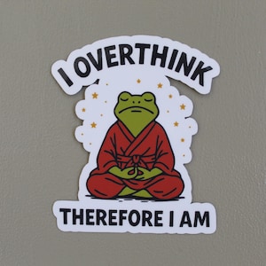 May include: A sticker featuring a green frog in a red robe, meditating with closed eyes. The text "I OVERTHINK" arches above the frog, and "THEREFORE I AM" is below. Gold stars surround the frog.