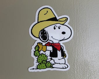 May include: A white sticker featuring Snoopy dressed as a cowboy, wearing a yellow hat, red bandana, black vest, and yellow gloves. He is holding a green cactus. The sticker has a white border.