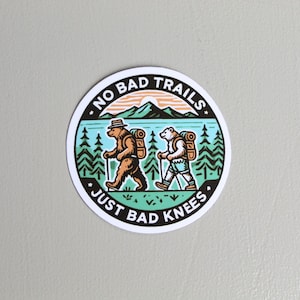 May include: A round sticker with a black border and white background. It features a cartoon illustration of a bear and a polar bear hiking with backpacks. The text reads "No Bad Trails, Just Bad Knees."